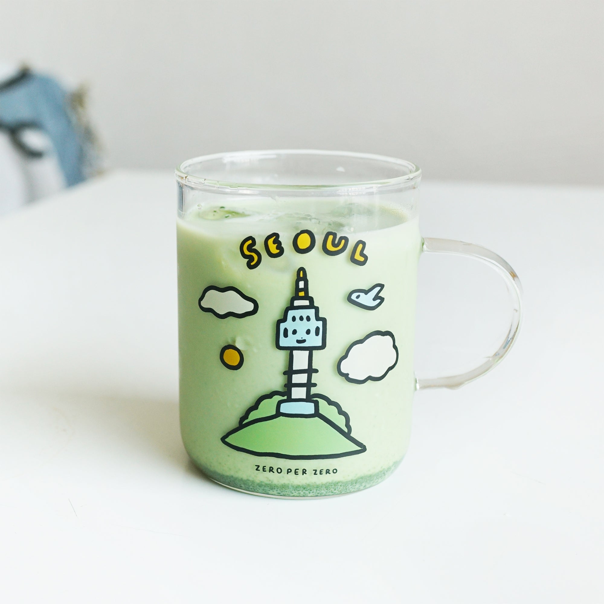 A glass drinking cup with matcha inside with a handle that has a colorful print of the N Seoul tower on it, created by Korean brand Zero Per Zero and for sale at Shim House